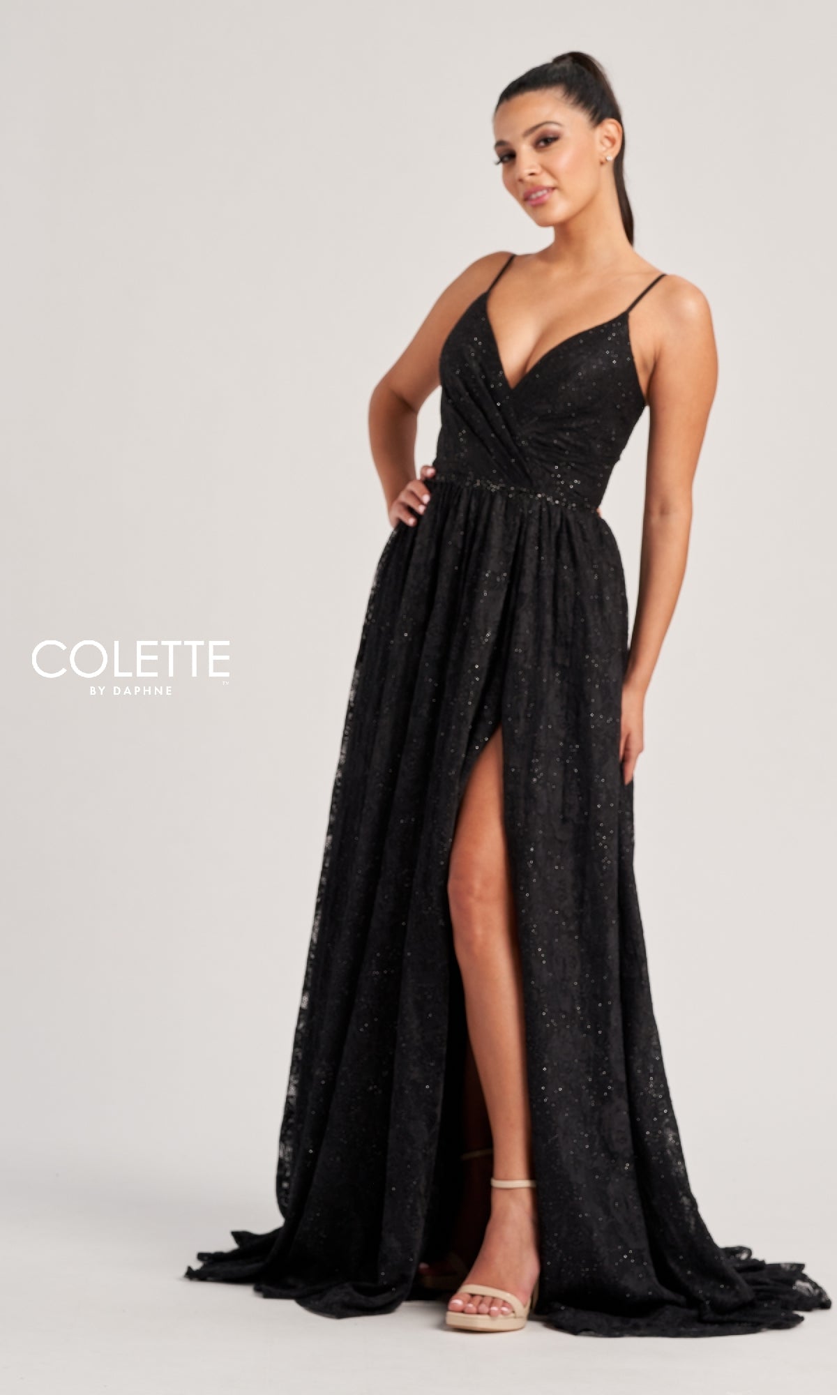 Woman wearing the Colette CL8010 long prom dress—sleeveless, floor-length, black with subtle sparkle and a thigh-high front slit—standing against a plain light background.